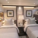 4d28cee4-9ba3-4708-8c21-2a473372107a - https://www.andyquinones.net Httpswww Andyquinones Net luxurious yacht cabin with two single beds soft gray bedding and matching pillows facing each other across a nightstand A mirror lamps framed art and decorative objects line the wall behind the beds porthole windows dot the opposite wall