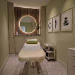 Massage therapy room interior design - https://www.andyquinones.net Httpswww Andyquinones Net treatment room with a massage bed round backlit mirror and beige cabinetry